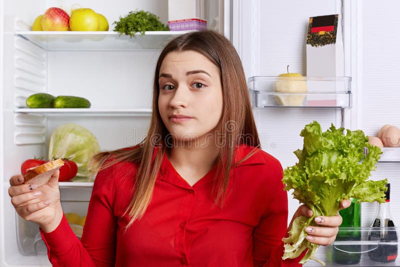 El modelo femenino joven vacilante sostiene lechuga y el bocadillo en manos, ` t del doesn conoce qué elegir, puede el ` t guarda foto de archivo El modelo femenino joven vacilante sostiene lechuga y el bocadillo en manos, ` t del doesn conoce qué elegir, puede el ` t guarda foto de archivo