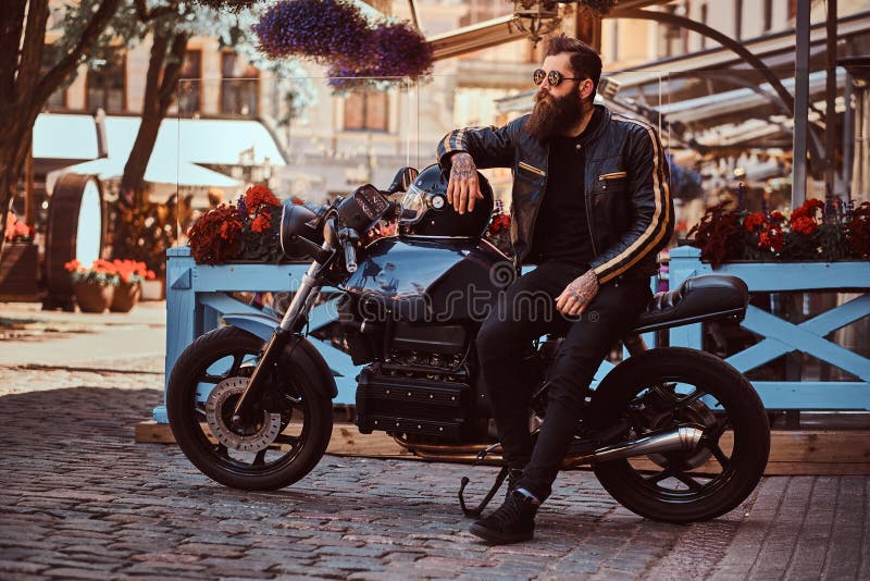 El motorista de moda elegante en gafas de sol se vistió en una chaqueta de cuero negra, sentándose en su motocicleta retra por en fotografía de archivo libre de regalías El motorista de moda elegante en gafas de sol se vistió en una chaqueta de cuero negra, sentándose en su motocicleta retra por en fotografía de archivo libre de regalías