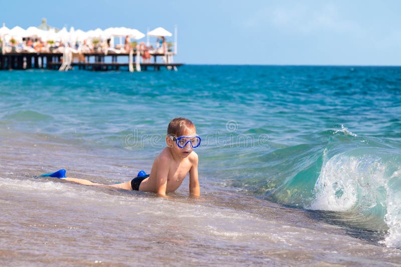 El muchacho caucásico en una tira de la resaca en la costa en una máscara y un tubo, toma el sol y espera una onda grande en vera foto de archivo libre de regalías El muchacho caucásico en una tira de la resaca en la costa en una máscara y un tubo, toma el sol y espera una onda grande en vera foto de archivo libre de regalías