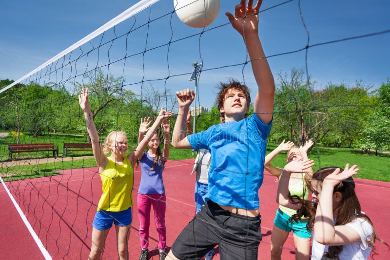 Jumping boy trying to catch ball with arms up plays volleyball with other teens near net on the playing court during sunny summer day. Jumping boy trying to catch ball with arms up plays volleyball with other teens near net on the playing court during sunny summer day