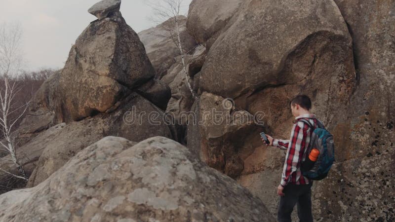 El muchacho joven en un equipo casual elegante y con la mochila turística vaga en las montañas, para y toma un selfie