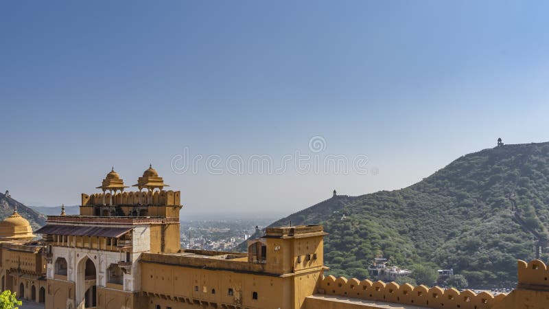The wall of the ancient Amber fort against the blue sky. The arched entrance, towers, windows are visible. In the distance, on the crest of the hill, the fortress defensive wall is visible. India. The wall of the ancient Amber fort against the blue sky. The arched entrance, towers, windows are visible. In the distance, on the crest of the hill, the fortress defensive wall is visible. India.