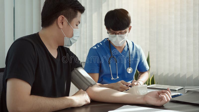 El médico asiático está utilizando un monitor de la presión arterial de los pacientes en el momento de su revisión anual y explica imagenes de archivo