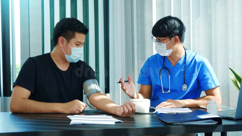 El médico asiático está utilizando un monitor de la presión arterial de los pacientes en el momento de su revisión anual y explica foto de archivo