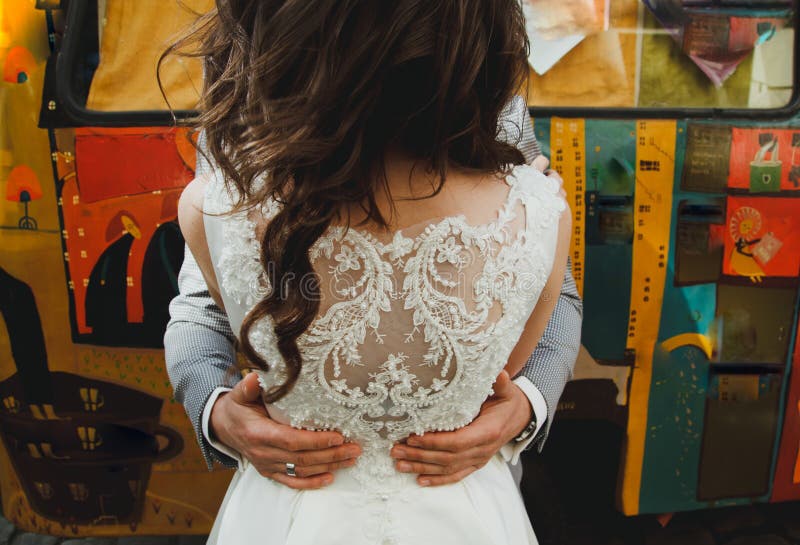 El novio está abrazando a la novia en vestido blanco elegante del satén con el cordón bordado detrás Autobús colorido del hippie imagen de archivo libre de regalías El novio está abrazando a la novia en vestido blanco elegante del satén con el cordón bordado detrás Autobús colorido del hippie imagen de archivo libre de regalías