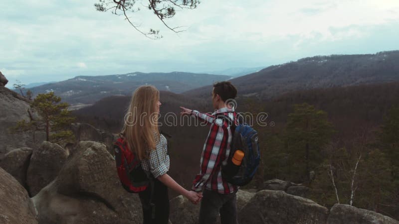 El par joven que sube las colinas rocosas, hombre joven elegante con una mochila da una mano a su novia hermosa con a