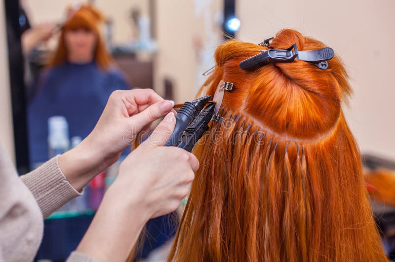 El peluquero hace extensiones del pelo a una muchacha joven, pelirroja, en un salón de belleza