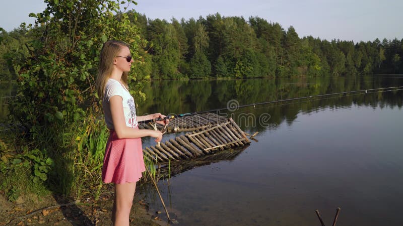 El pescador rubio de la chica joven está pescando en el lago del bosque, llevando a cabo las torsiones de la caña de pescar la bo almacen de video