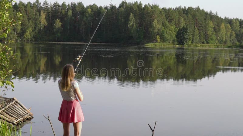 El pescador rubio de la chica joven está pescando en el lago del bosque La mujer atractiva en vestido corto aprende pescar en el almacen de video