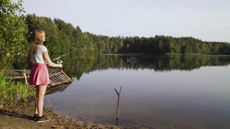 El pescador rubio de la chica joven está pescando en el lago del bosque La mujer atractiva en vestido corto aprende pescar en el almacen de video