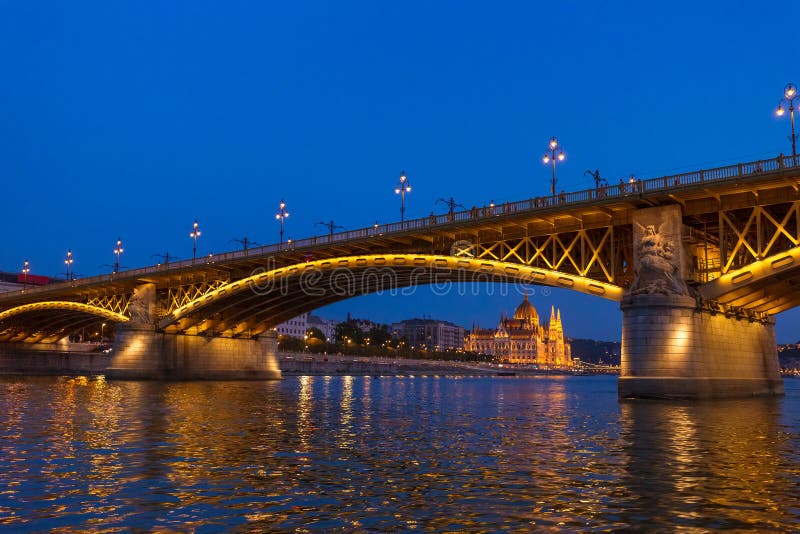 The Margaret Bridge with Parliament Building on background in Budapest, Hungary, illuminated above the Danube river at night. The Margaret Bridge with Parliament Building on background in Budapest, Hungary, illuminated above the Danube river at night