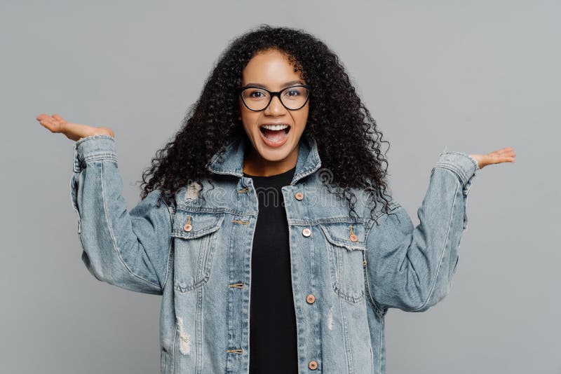 El retrato de la mujer afroamericana rizada satisfecha alegre aumenta las palmas, finge llevar a cabo algo, lleva los vidrios ópt fotos de archivo libres de regalías El retrato de la mujer afroamericana rizada satisfecha alegre aumenta las palmas, finge llevar a cabo algo, lleva los vidrios ópt fotos de archivo libres de regalías