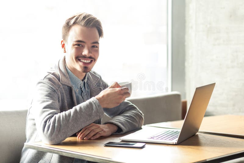 El retrato del hombre de negocios joven barbudo feliz hermoso en chaqueta gris se está sentando en café y tener una rotura con la fotografía de archivo libre de regalías El retrato del hombre de negocios joven barbudo feliz hermoso en chaqueta gris se está sentando en café y tener una rotura con la fotografía de archivo libre de regalías