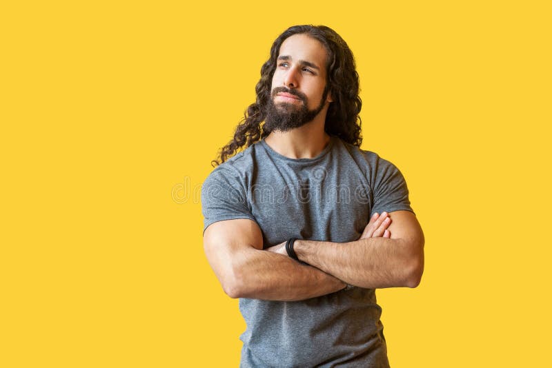 El retrato del hombre joven barbudo pensativo con el pelo rizado largo en la situación gris de la camiseta, cruzó las manos, mira fotos de archivo libres de regalías El retrato del hombre joven barbudo pensativo con el pelo rizado largo en la situación gris de la camiseta, cruzó las manos, mira fotos de archivo libres de regalías