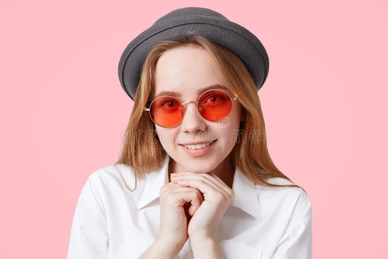 El retrato del modelo femenino encantado feliz en gafas de sol rojas redondas y sombrero elegante, guarda las manos debajo de la fotografía de archivo El retrato del modelo femenino encantado feliz en gafas de sol rojas redondas y sombrero elegante, guarda las manos debajo de la fotografía de archivo