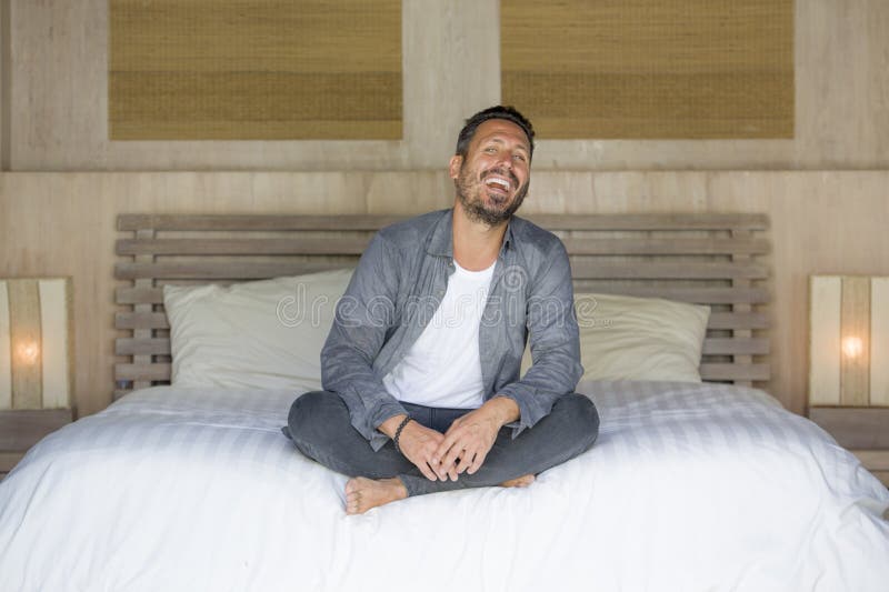 El retrato interior de 30s al hombre feliz y hermoso de 40s en casa en la camisa sport y los vaqueros que se sentaban en cama rel fotos de archivo libres de regalías El retrato interior de 30s al hombre feliz y hermoso de 40s en casa en la camisa sport y los vaqueros que se sentaban en cama rel fotos de archivo libres de regalías