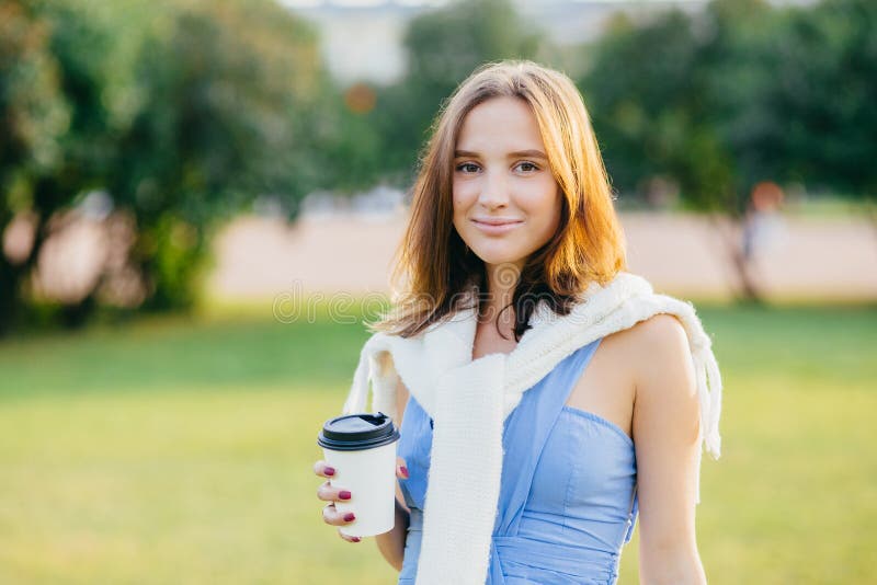 El tiro al aire libre de la hembra europea joven de mirada agradable con el pelo oscuro, café para llevar de los controles, pasa fotografía de archivo libre de regalías El tiro al aire libre de la hembra europea joven de mirada agradable con el pelo oscuro, café para llevar de los controles, pasa fotografía de archivo libre de regalías