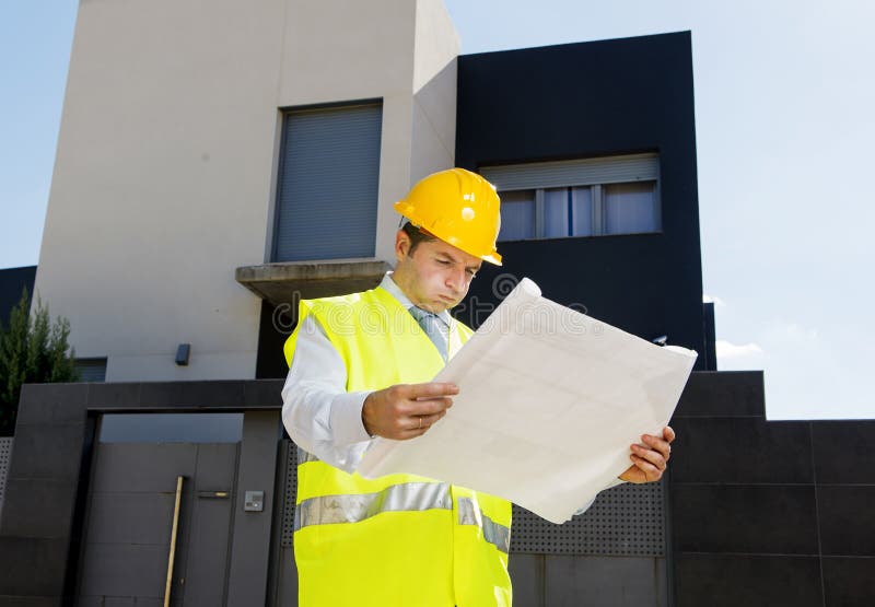 Young attractive foreman worker in stress supervising building blueprints outdoors wearing construction helmet and vest in housing development and real estate concept. Young attractive foreman worker in stress supervising building blueprints outdoors wearing construction helmet and vest in housing development and real estate concept