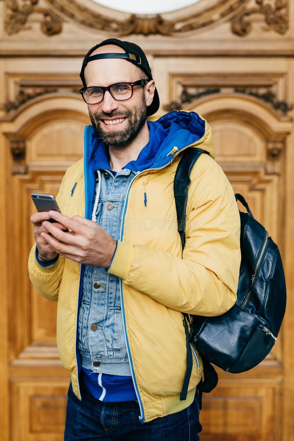 El turista del inconformista en los vidrios, casquillo y mochila amarilla y smartphone de la tenencia del anorak teniendo excursi foto de archivo El turista del inconformista en los vidrios, casquillo y mochila amarilla y smartphone de la tenencia del anorak teniendo excursi foto de archivo
