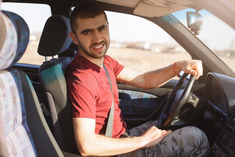 El varón joven sin afeitar hermoso con rastrojo se sienta en coche, tiene rotura después de conducir, mira la cámara, disfruta de fotografía de archivo libre de regalías El varón joven sin afeitar hermoso con rastrojo se sienta en coche, tiene rotura después de conducir, mira la cámara, disfruta de fotografía de archivo libre de regalías