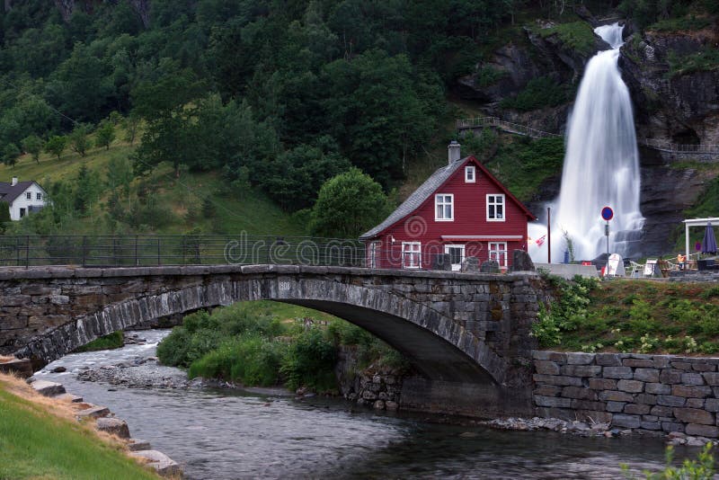 El viaje a Noruega, una casa brillante se coloca cerca de una cascada, que fluye en un río, a través del cual se construye un pue fotos de archivo El viaje a Noruega, una casa brillante se coloca cerca de una cascada, que fluye en un río, a través del cual se construye un pue fotos de archivo