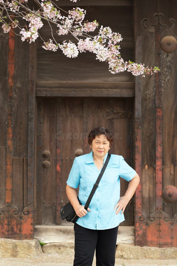 Elder asian woman standing with hand on the hips with smile on camera in Himeji castle area in cherry blossom sakura period in Japan.