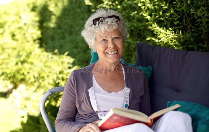 Elder woman sitting on a chair in backyard garden holding a book and looking at camera smiling. Elder woman sitting on a chair in backyard garden holding a book and looking at camera smiling