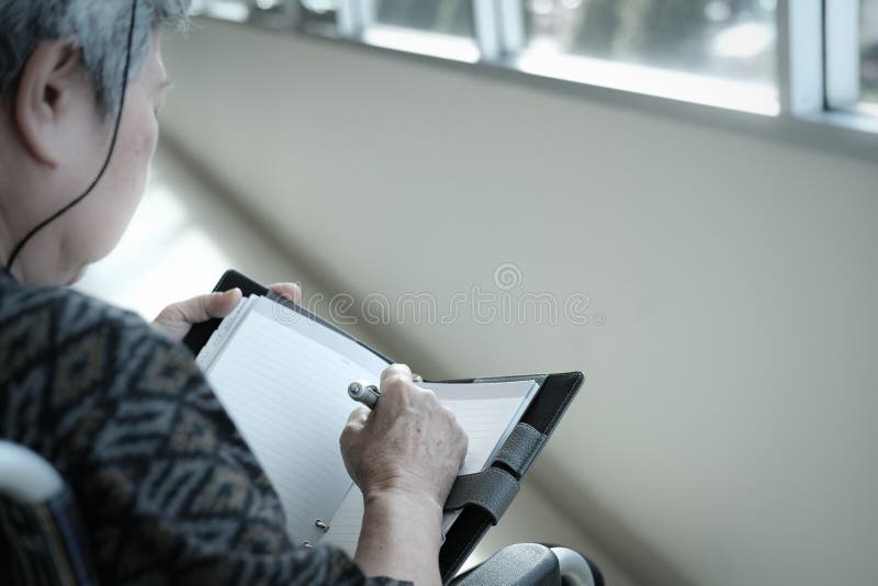 elder woman on wheelchair writing note on notebook near window.