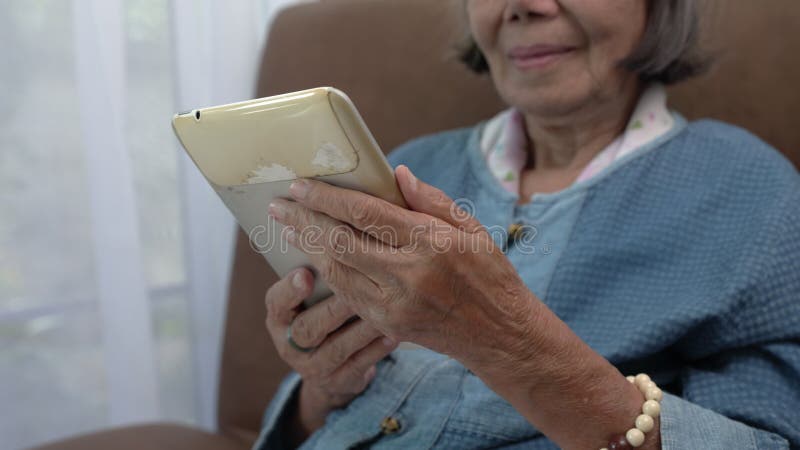 Elderly Asian woman relax on couch in living room browsing wireless Internet on obsolete tablet