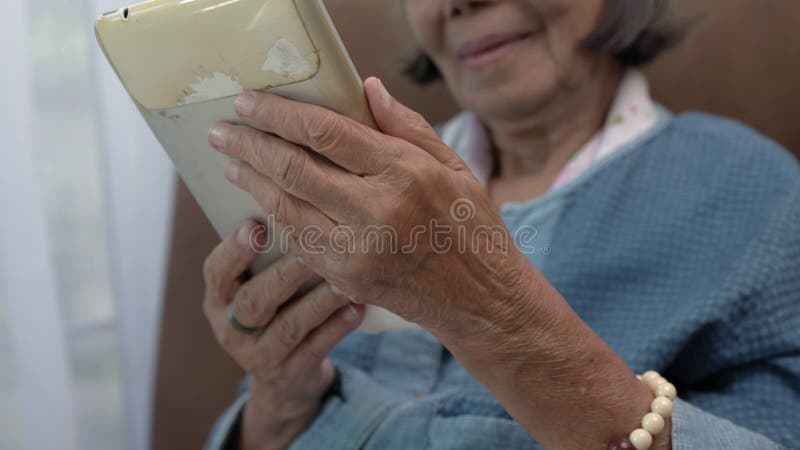 Elderly asian woman relax on couch in living room browsing wireless Internet on obsolete tablet