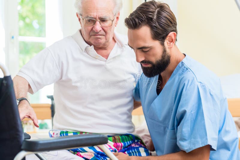 Elderly care nurse helping senior from bed to wheel chair in hospital or nursing home