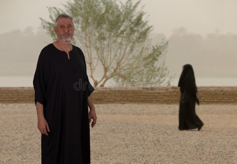 An elderly Egyptian in black robe stands on the bank of the Nile. A woman walks in the background.