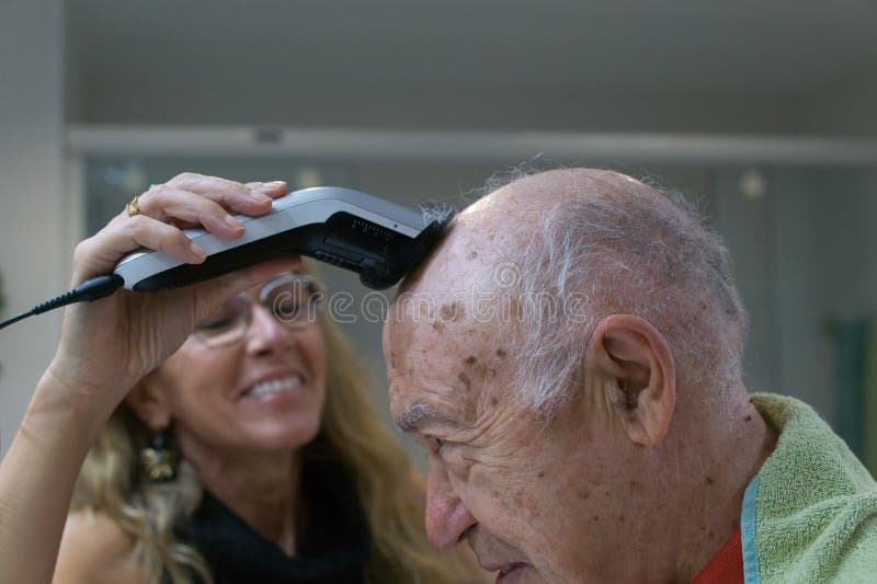 Elderly man having his head shaved by a smiling woman in a casual setting, showcasing care stock photos