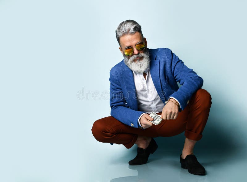 Elderly man in white shirt, jacket, brown pants, black loafers and sunglasses. Squatting, showing cash, posing on blue background. Bearded elderly man in a white royalty free stock photos Elderly man in white shirt, jacket, brown pants, black loafers and sunglasses. Squatting, showing cash, posing on blue background. Bearded elderly man in a white royalty free stock photos