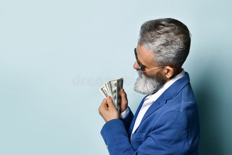 Elderly man in white shirt, jacket, brown pants and sunglasses. He showing fan of hundred dollar bills, posing on blue background. Bearded elderly man in a white stock image Elderly man in white shirt, jacket, brown pants and sunglasses. He showing fan of hundred dollar bills, posing on blue background. Bearded elderly man in a white stock image