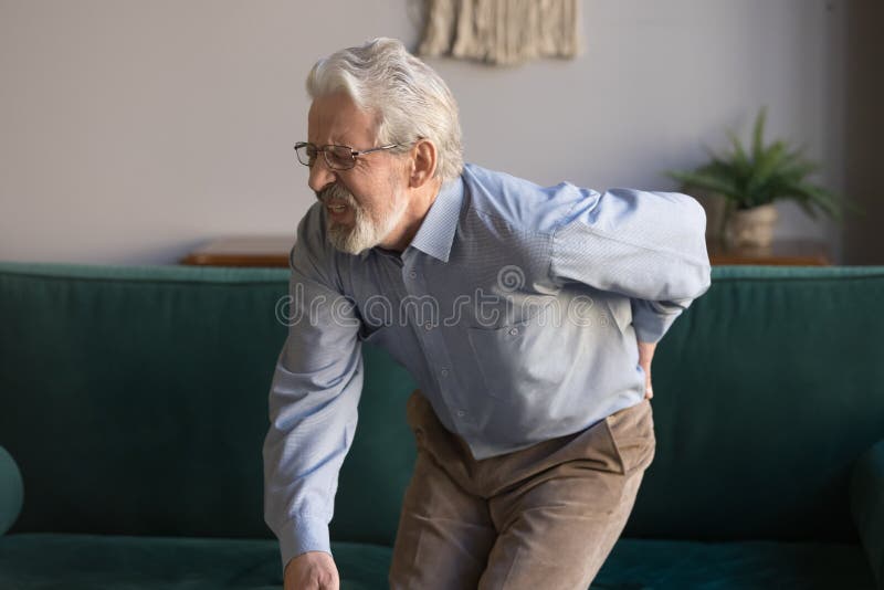 Aged grey haired sixty years man in glasses writhes in pain suffers from low back strain, touch rubbing or massaging loin reduces backache. Degenerative disk disease, pinched nerve rheumatism concept. Aged grey haired sixty years man in glasses writhes in pain suffers from low back strain, touch rubbing or massaging loin reduces backache. Degenerative disk disease, pinched nerve rheumatism concept