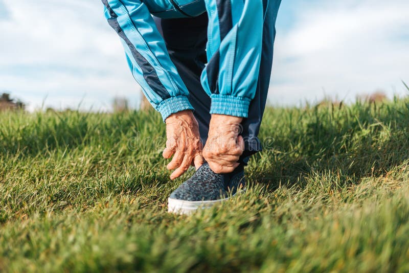 An elderly woman adjusts her sports shoes on her foot while standing on the grass. Close-up. The concept of an active. Lifestyle and health problems stock images