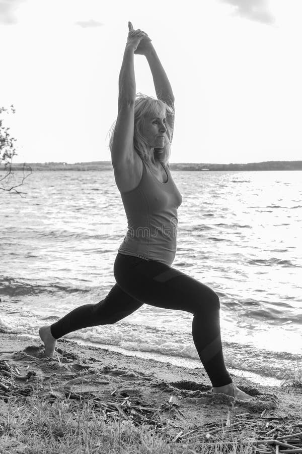 An elderly woman is doing sports on the lawn on the shore of the lake. The woman stands in the warrior pose with her hands up. Yoga pose. Outdoor sports