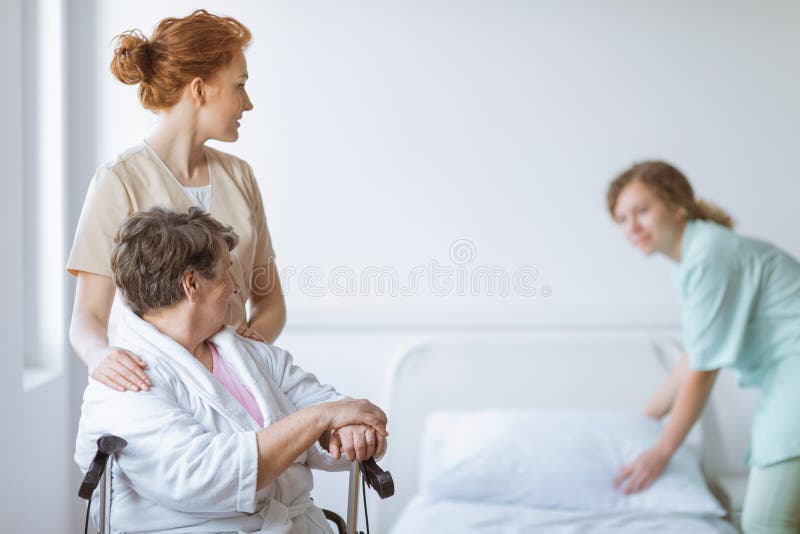 Elderly woman on wheelchair in nursing home with helpful doctor at her side and young nurse making the bed royalty free stock photos