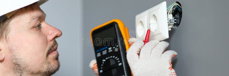 Electrician in helmet measures level electricity. Combined electrical measuring instrument, combining several functions. Handheld multimeter for basic measurements and troubleshooting at outlet