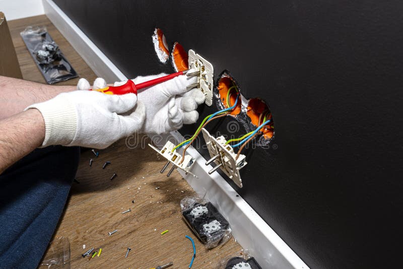 An electrician uses a red screwdriver to screw a double grounded electrical socket in a row to the electrical box in the black wal