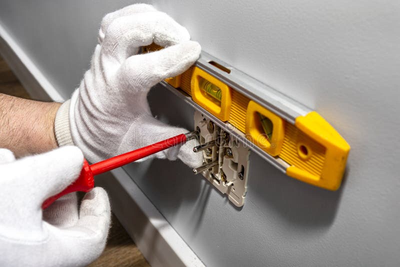 An electrician uses a red screwdriver to screw a double grounded electrical outlet into the wall of the room and use a spirit leve
