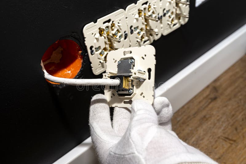 An electrician in white gloves is installing a TV socket and satellite connector in the black wall of the room.