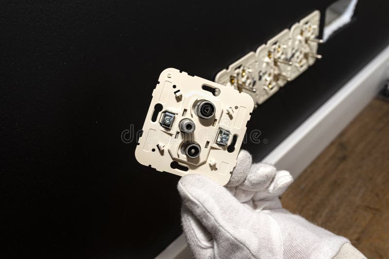 An electrician in white gloves is installing a TV socket and satellite connector in the black wall of the room.