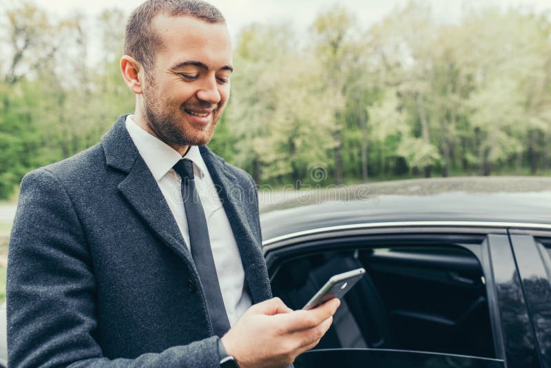 Elegancka poważna przystojna młoda Kaukaska osoba jest ubranym żakiet z samochodem, sprawdza emaila na telefonie komórkowym zdjęcia stock Elegancka poważna przystojna młoda Kaukaska osoba jest ubranym żakiet z samochodem, sprawdza emaila na telefonie komórkowym zdjęcia stock