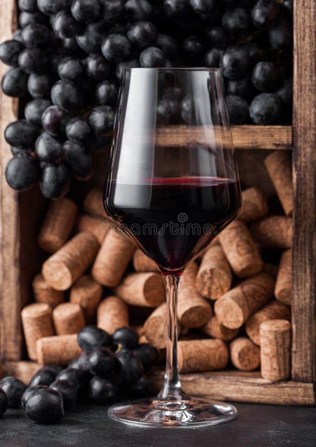 Elegant glass of red wine with dark grapes and corks inside vintage wooden box on black stone background. Elegant glass of red wine with dark grapes and corks inside vintage wooden box on black stone background