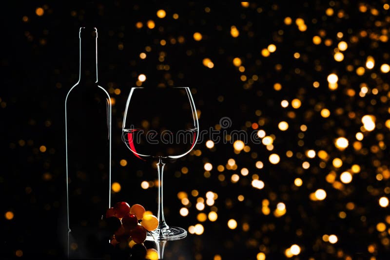 Elegant red wine glass, wine bottle and bunch of grapes on the dark background and blurred defocused lights as Christmas decorations background. Party concept. Valentine`s day, Birthday concept. Elegant red wine glass, wine bottle and bunch of grapes on the dark background and blurred defocused lights as Christmas decorations background. Party concept. Valentine`s day, Birthday concept.