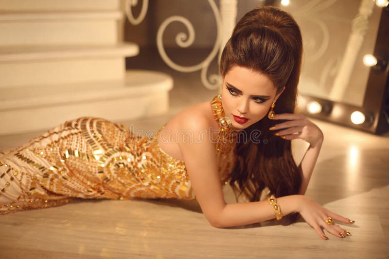 Elegant fashion brunette woman in luxury golden dress. Hair style and red lips Makeup. Manicured nails. girl model lying in glamour gown in modern interior.