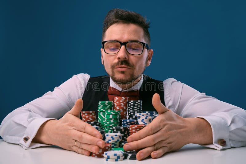 Man in glasses, black vest and shirt sitting at white table with stacks of chips on it, posing on blue background stock photo