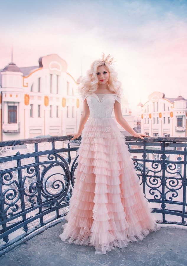 Beautiful elegant princess woman. Queen lady standing on balcony. Backdrop art retro old cityscape blue sky. Long pink medieval vintage dress. Blonde wavy hair volume wig. Royal gold crown cute face. Beautiful elegant princess woman. Queen lady standing on balcony. Backdrop art retro old cityscape blue sky. Long pink medieval vintage dress. Blonde wavy hair volume wig. Royal gold crown cute face